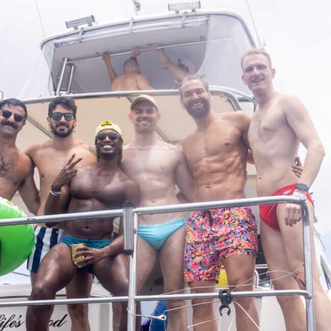 Six men in swimwear stand on a boat, smiling and posing for a photo. One holds an inflatable llama ring, another shows a peace sign. A sunny day with a clear sky is in the background.