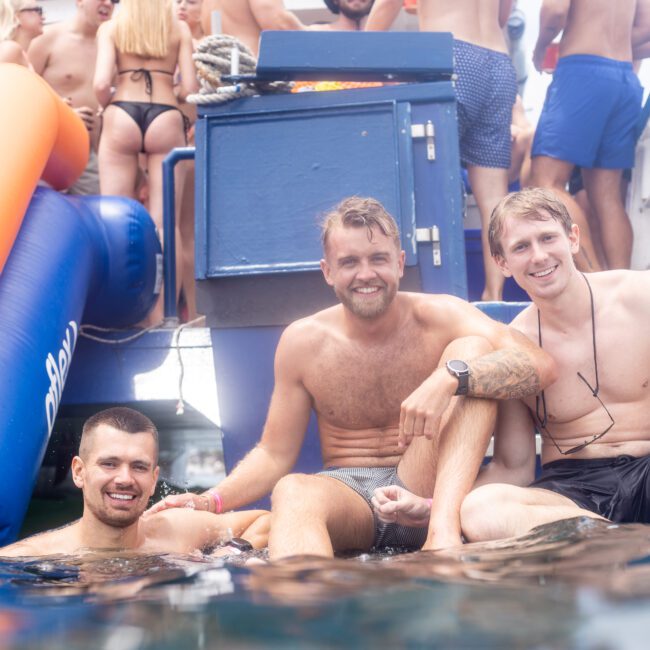 Three men sit at the edge of a boat, smiling and partially submerged in water. The boat is filled with other people enjoying a sunny day, with inflatables visible nearby. The atmosphere is lively and festive.