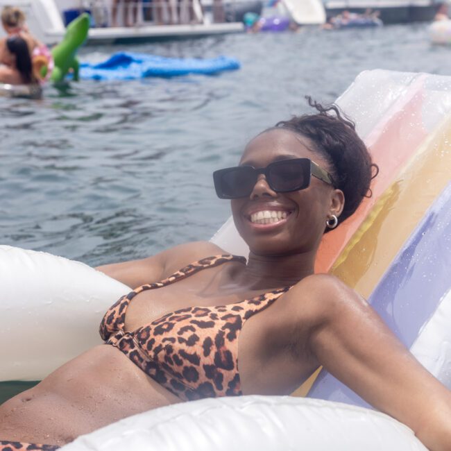 A woman is lounging on a float in the water, wearing a leopard print bikini and sunglasses, smiling at the camera. Other people are in the background enjoying the water.