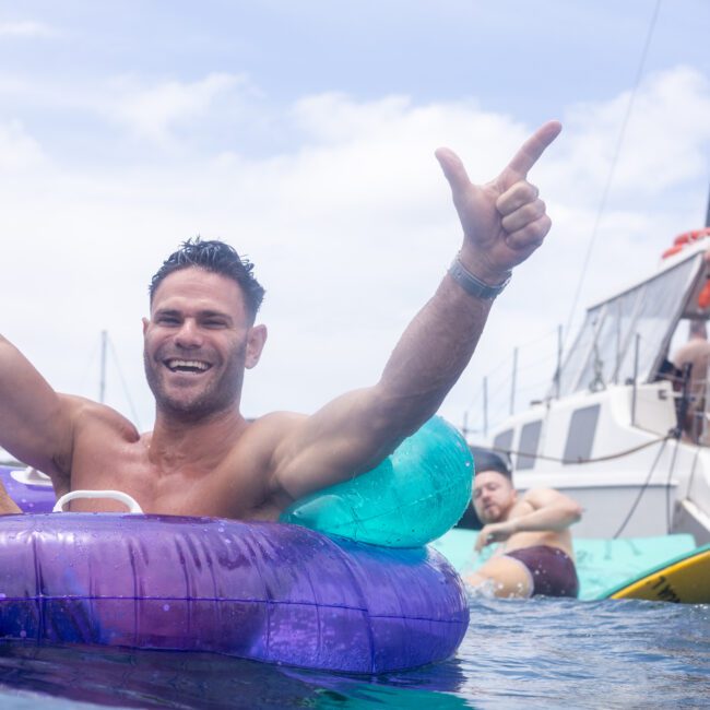 A man smiles while floating on a purple inflatable ring in the water, holding a drink and gesturing with both hands. In the background, others relax on a boat under a partly cloudy sky.