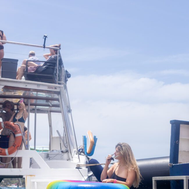 A woman in a swimsuit sits on a colorful inflatable ring in the water near a boat. She holds a drink while other people are on the boat, enjoying the sunny day. The boat is equipped with a diving platform.