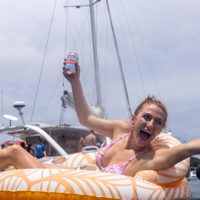 A woman in a bikini joyfully raises a can while lounging on an inflatable float in the water. A sailboat with people on board is in the background under a partly cloudy sky.