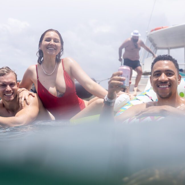 Three people smiling in the water by a yacht. One person is giving a peace sign, another is holding a drink in an inflatable ring. A few more people are visible on the yacht in the background. The scene is sunny and joyful.