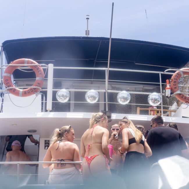 A group of people in swimwear stand on a yacht, gathered and socializing. Four lifebuoys are visible, and the ocean can be seen at the bottom of the image. The scene suggests a sunny day with clear skies.