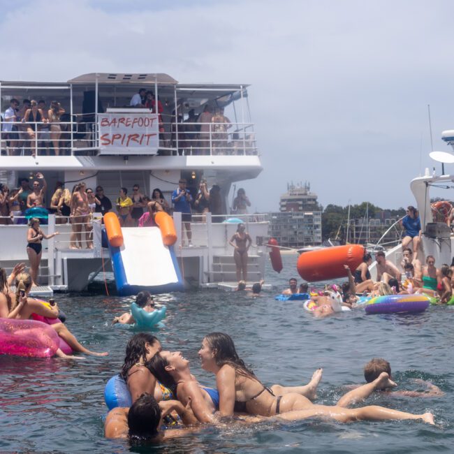 A lively scene of a boat party with people swimming and floating on inflatables in the water. A large boat labeled "Barefoot Spirit" and another smaller vessel are crowded with people enjoying the sunny day.