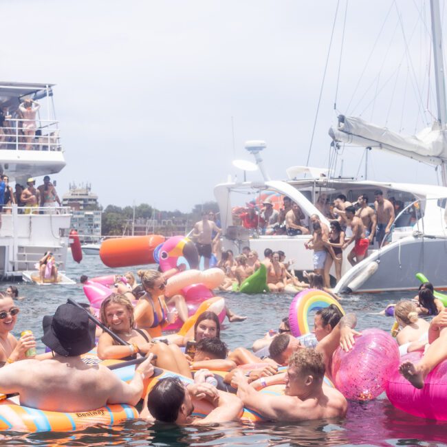 A vibrant boat party scene with numerous people on colorful inflatable floats in the water. Several boats are anchored nearby, including a yacht and a catamaran. People are socializing and enjoying the sunny weather.