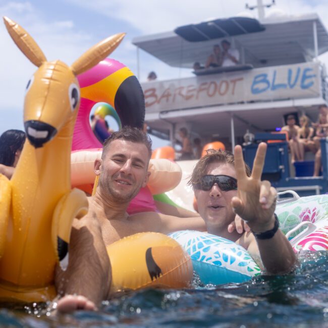 Two people relax on colorful inflatable floats in a body of water, with one person making a peace sign. A boat with "Barefoot Blue" written on it is in the background. The sky is clear and sunny.