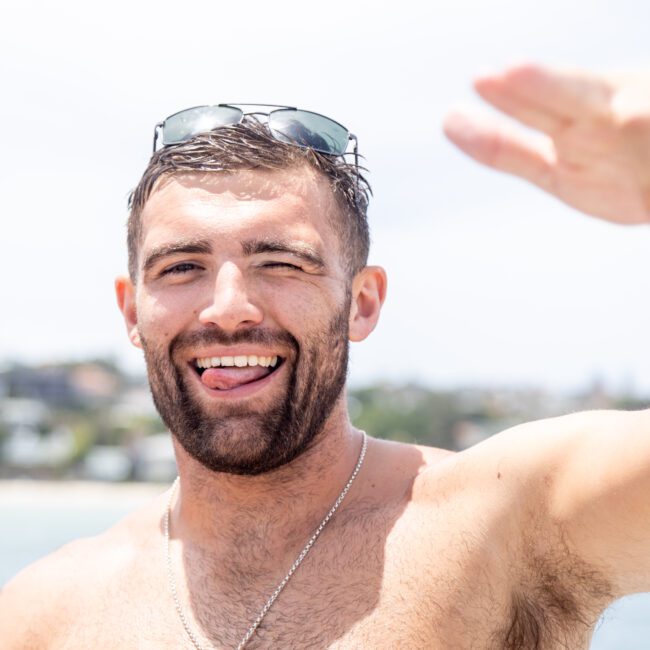 A person with short hair is smiling and sticking out their tongue while making a peace sign with one hand. They are wearing sunglasses on their head and a chain necklace. The background shows a beach and blurred buildings.