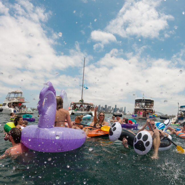 A group of people enjoying a day in the water, surrounded by colorful inflatable pool floats, including a purple unicorn and a black and white paw print float. Boats and a city skyline are visible in the background under a partly cloudy sky.