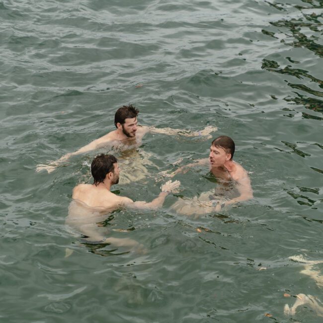 Three men are swimming in a body of water, engaging in conversation. The water is greenish with ripples around them, indicating movement. They appear relaxed and focused on each other.