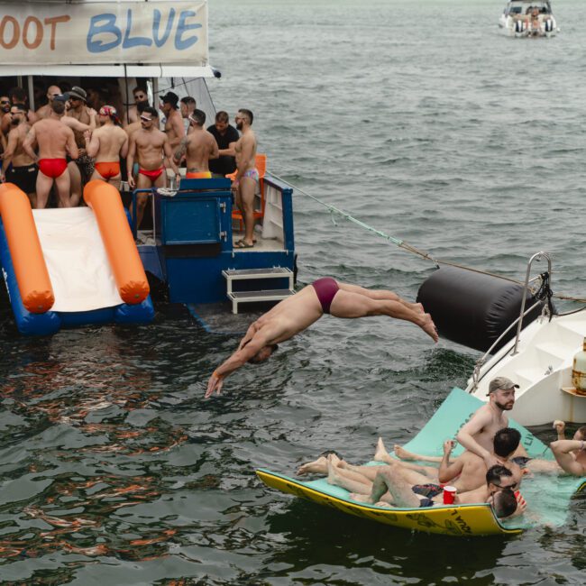 A man dives into the water from a boat where a group of people in red swimwear are gathered. Nearby, several people relax on a float, enjoying a day on the water. Trees and buildings are visible in the background.