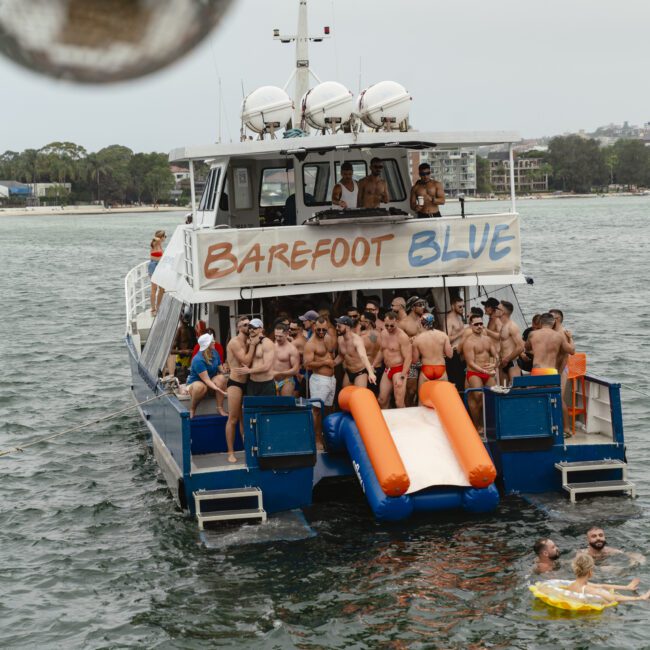 A boat named "Barefoot Blue" on the water is filled with people in swimsuits. Two orange and blue inflatable slides extend into the water. Several people are swimming nearby. The background shows a shoreline with trees and buildings.