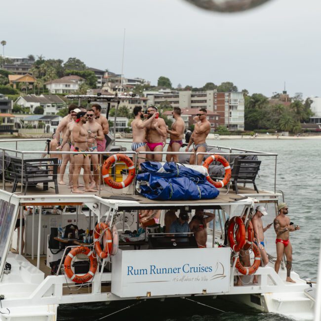 A group of people in swimwear enjoying a party on the upper deck of a Rum Runner Cruises boat. They are socializing and dancing with a backdrop of waterfront buildings and greenery. Life preservers and a blue tarp are visible on the boat.