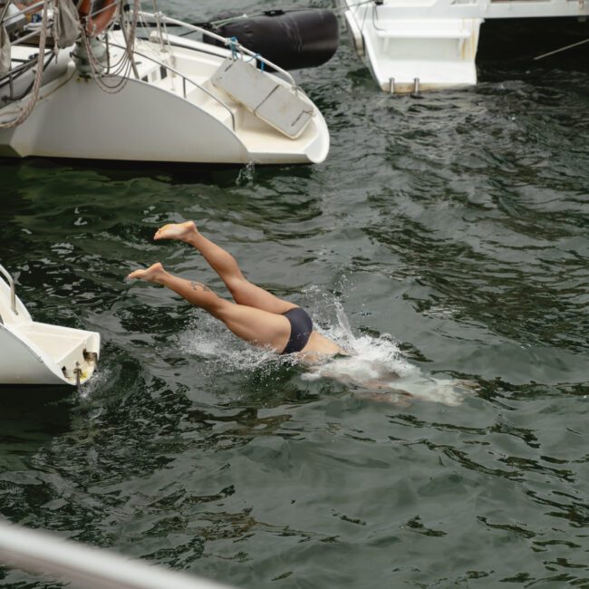 A person diving into the water from a boat, surrounded by other boats. The scene captures the splash and movement of the dive, with calm water and overcast skies.