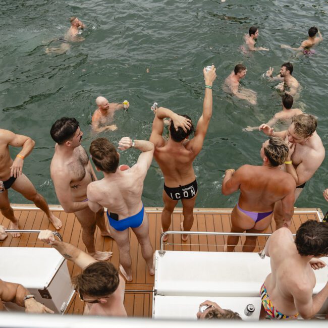 A group of people in swimwear enjoying a party on a boat and swimming in the water. Some are dancing and holding drinks, while others are in the water or sitting on the boat, creating a lively and festive atmosphere.