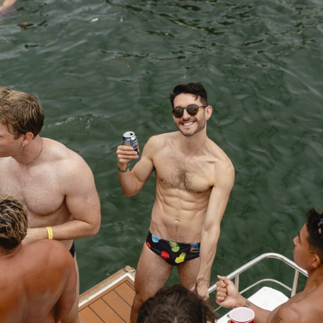 A shirtless man in swim trunks smiles while holding a beverage on a boat deck. He is surrounded by friends who are also in swimwear. The group appears to be enjoying a sunny day, with water in the background.