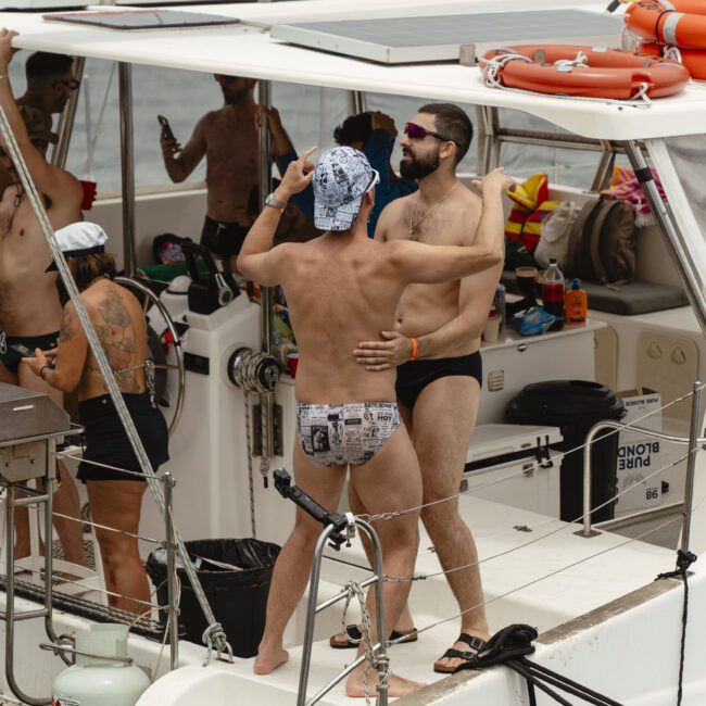 Two people in swimwear dance on the deck of a boat. They are surrounded by others enjoying the boat ride, with some taking photos and socializing. The scene is lively and relaxed, set against a backdrop of water and sky.