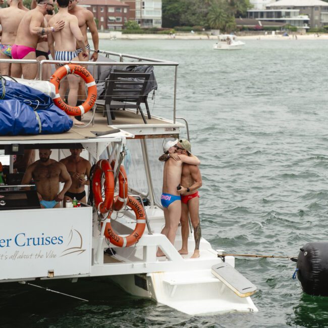 A group of people in swimwear socialize on a boat in a body of water. Two individuals hug near the boat's edge. Buildings and trees are visible on the shore in the background.