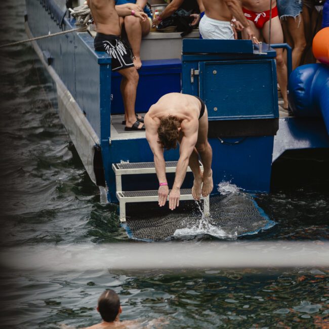 A group of people are having fun on a boat. A man is diving into the water, while another person is swimming nearby. Others on the boat are watching and relaxing. The scene is lively and set on a sunny day.