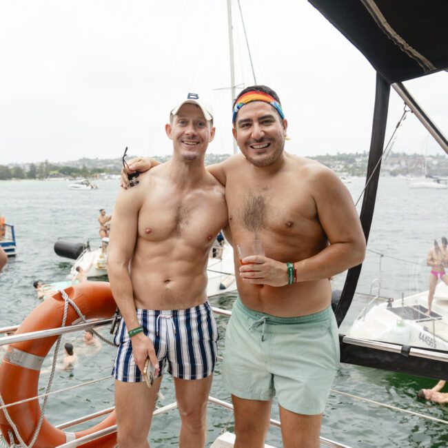 Two men in swim trunks stand smiling on a boat deck, with one arm around each other. They are surrounded by water and other boats. The sky is overcast, and people swim in the background. A lifesaver is visible on the left.