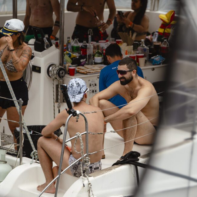 A group of people relax on a boat. Some are sitting and chatting, while others stand by a table with drinks and snacks. The scene is casual, with everyone wearing swimwear. The boat's equipment and railing are visible in the foreground.