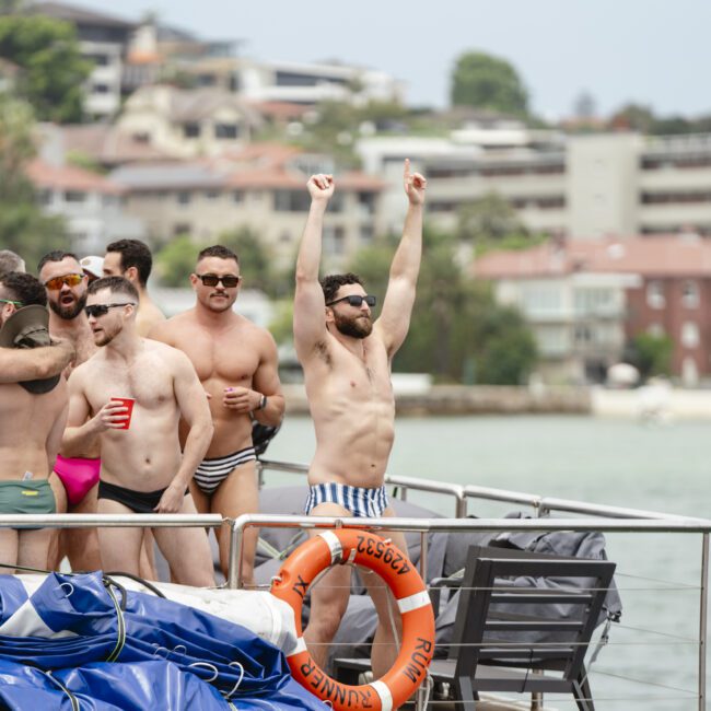 A group of men in swimwear enjoy a lively boat party near the shore. One man raises his arms in excitement, while others chat and hold drinks. Life preservers are visible, and buildings can be seen in the background.