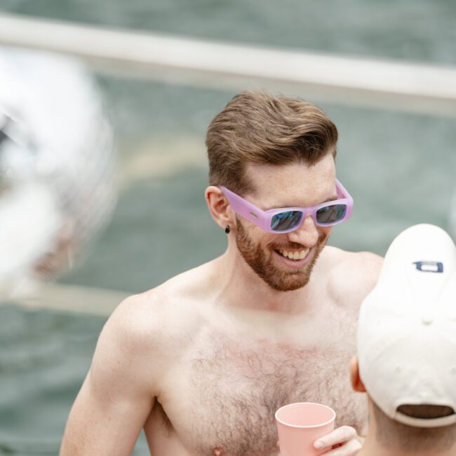 A man with short brown hair and a beard, wearing purple sunglasses and a silver earring, smiles holding a pink cup. He's shirtless and talking to another person wearing a white cap. The scene is outdoors, likely near water.