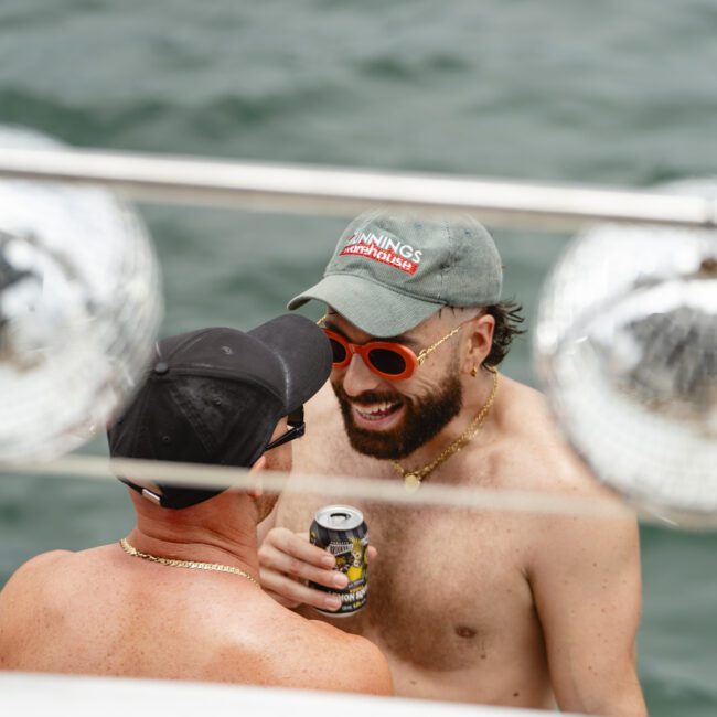 Two men wearing caps and sunglasses are smiling and chatting on a boat. One is holding a can of drink. They appear to be shirtless and are enjoying a relaxed moment on the water.