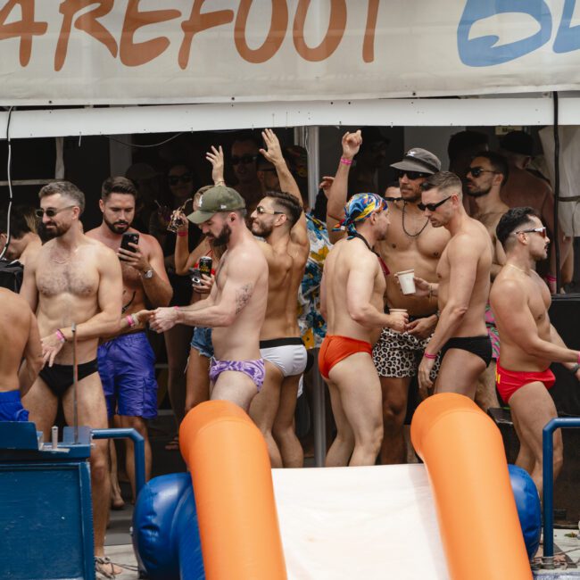 A lively group of men in colorful swimwear enjoy a party on a boat. Some are dancing, others are chatting or holding drinks. The boat features a slide and has a banner with partial text visible.