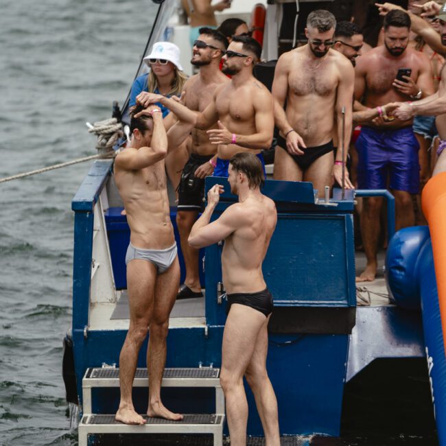 A group of people in swimwear gathers on the back of a boat. Some are standing in the water, while others watch and take photos. The scene suggests a fun, social event on the water.