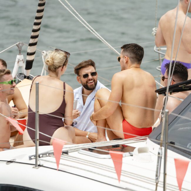 A group of people in swimsuits sit and chat on a sailboat. The boat is adorned with triangular flags, and the water is visible in the background. The atmosphere is casual and sunny.