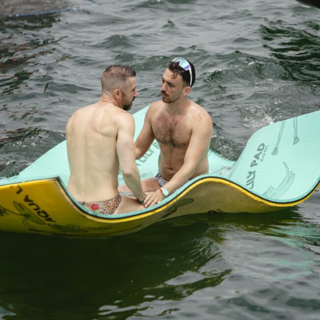 Two men sitting on a floating mat on a lake. Both are shirtless, wearing swim trunks, and facing each other. One person has sunglasses on their head. The water is calm, with a dock visible in the background.