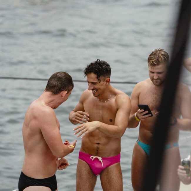 Three people in swimwear stand and chat on a boat. One is holding a phone, another is applying lotion. The water and another boat are visible in the background.