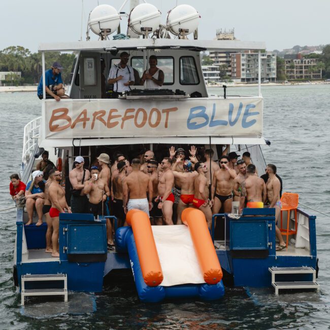 A group of people in swimsuits stands on a boat named "Barefoot Blue." They are enjoying a sunny day on the water, with an inflatable slide at the rear. Some people are raising their hands and smiling. Buildings are visible in the background.