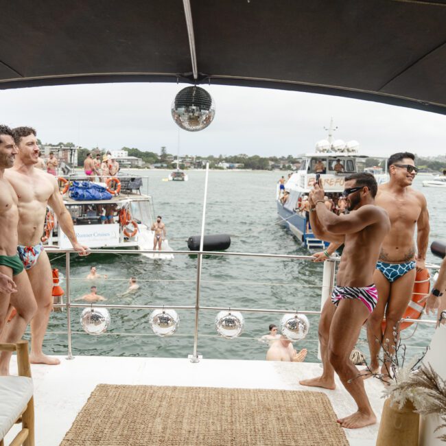 A group of people in colorful swimwear are on a boat taking photos. Other boats and swimmers are visible in the water. The scene is lively with clear skies and calm waters.