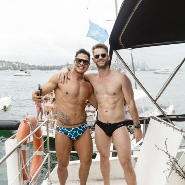 Two men in swimwear stand side by side on a boat, smiling with an arm around each other. In the background, people are on another boat with a blurred city skyline and ocean visible.