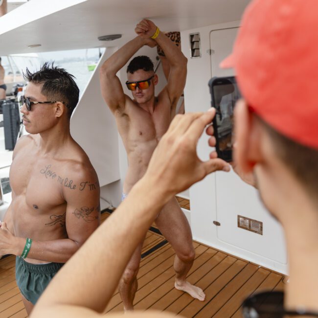 A group of shirtless men on a boat deck. One man is striking a pose wearing sunglasses, while another man in a red cap takes a photo with a smartphone. They appear to be enjoying a sunny day on the water.