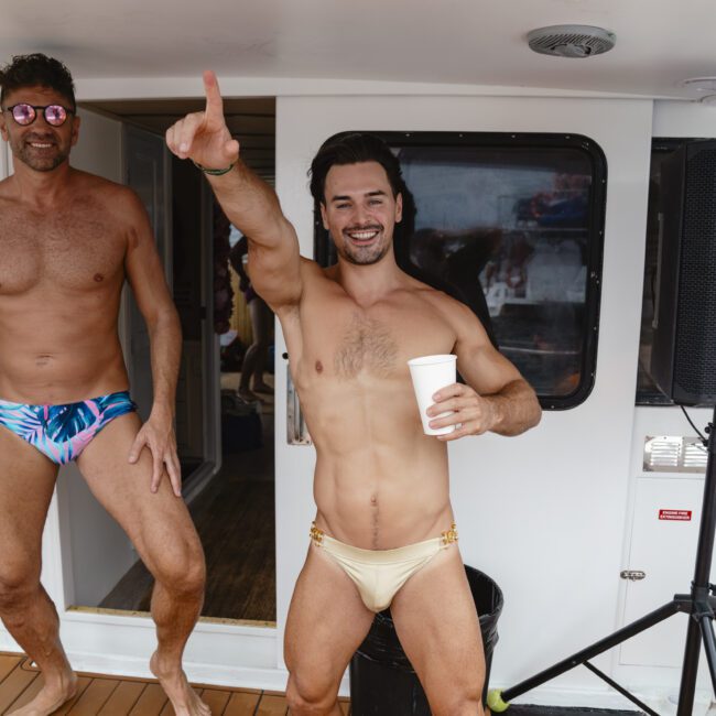 Two men in swim briefs are having fun on a boat. One man is pointing and holding a cup, while the other is smiling and wearing colorful sunglasses. The scene suggests a joyful, relaxed atmosphere on the boat deck.