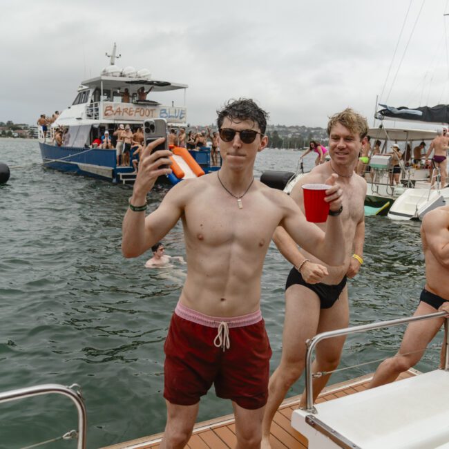 A man in red swim trunks and sunglasses holds a phone and a red cup on a boat. Other people in swimsuits are visible in the background. The scene is festive, with boats and water in the background.