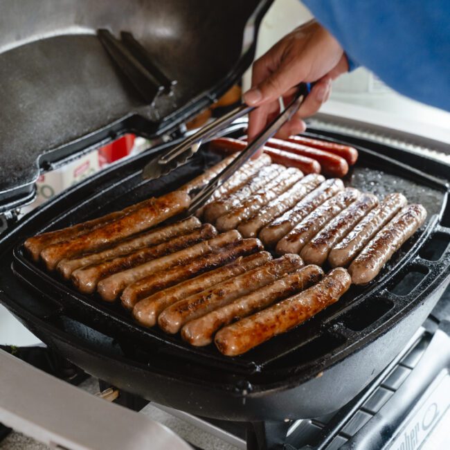 A person grills multiple sausages on a barbecue grill with the lid open. The sausages are evenly spaced and being turned with tongs, showcasing a delicious outdoor cooking scene.