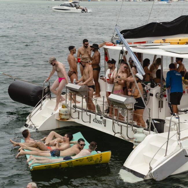 A group of people are enjoying a sunny day on a boat and in the water. Some are lounging on a floating mat, while others stand or sit on the boat. The background features a calm body of water with a few distant boats.