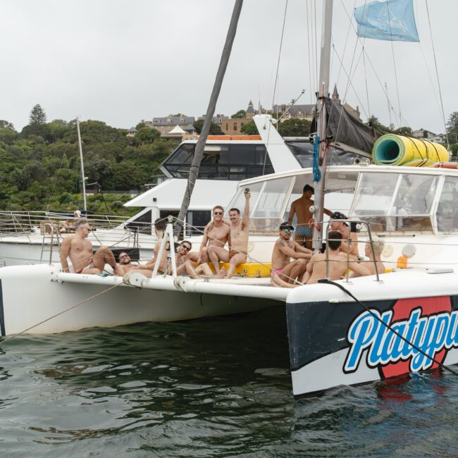 A group of people are relaxing on a white catamaran named "Platypus" in a calm body of water. They are wearing swimwear and appear to be enjoying a leisurely day. Trees and houses are visible in the background under an overcast sky.