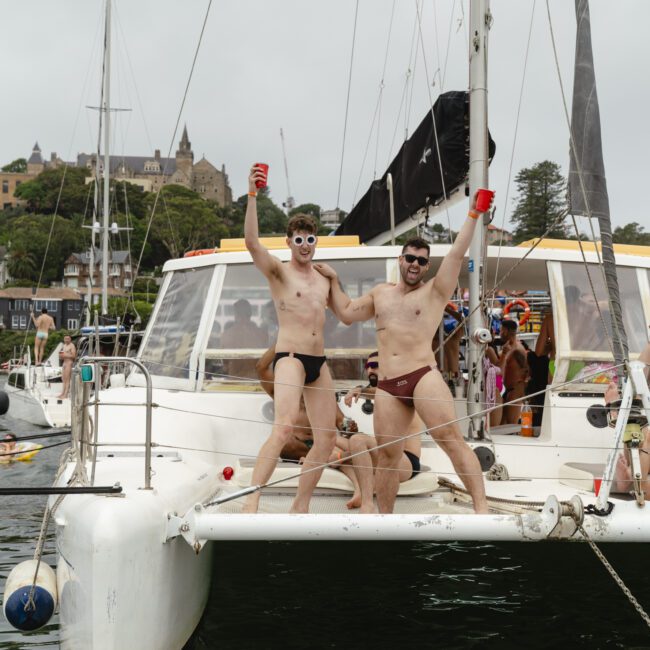 Two people in swimwear stand on the deck of a catamaran, holding red cups and cheering. The boat is docked, with buildings and trees visible in the background. It's a lively scene, suggesting a celebratory or festive atmosphere.