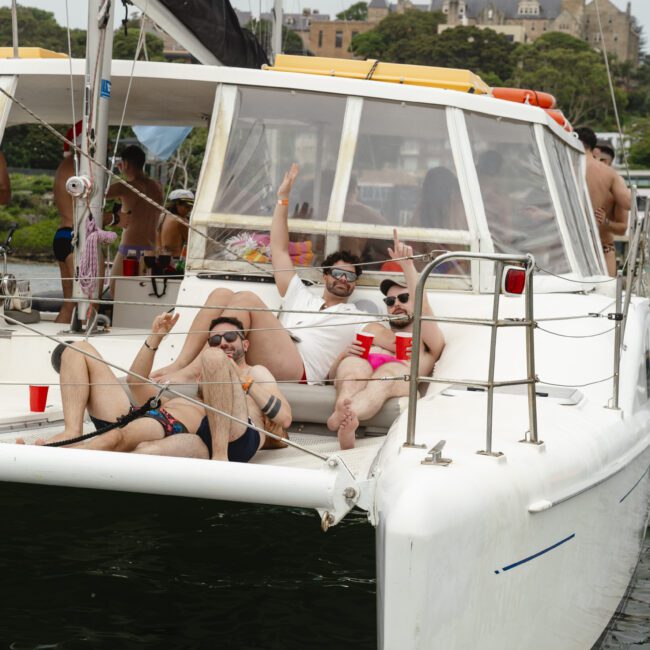 A group of people relax on a catamaran, enjoying drinks and lounging in swimwear. The boat is anchored on a body of water, and greenery and buildings line the shore in the background. The atmosphere is festive and leisurely.