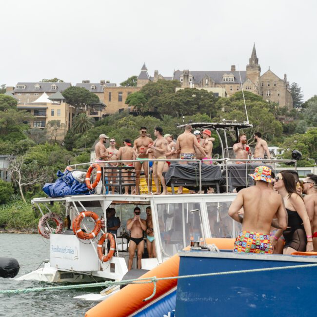 A group of people in swimsuits are enjoying a party on a large boat. The boat is on a body of water with buildings and a hill in the background. Inflatable water toys float nearby, and the scene is festive and lively.