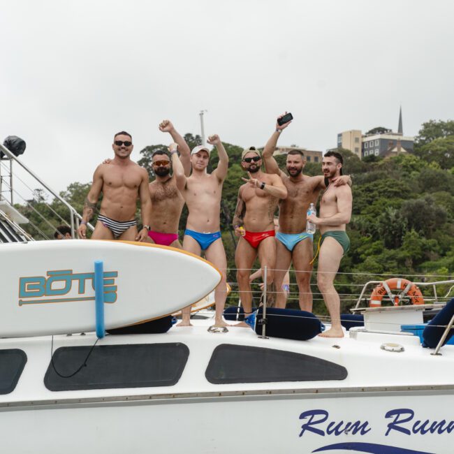 A group of six men in swimsuits stand cheerful and waving on a boat named "Rum Runner" on a cloudy day. The boat is in the water with a cityscape and trees in the background.