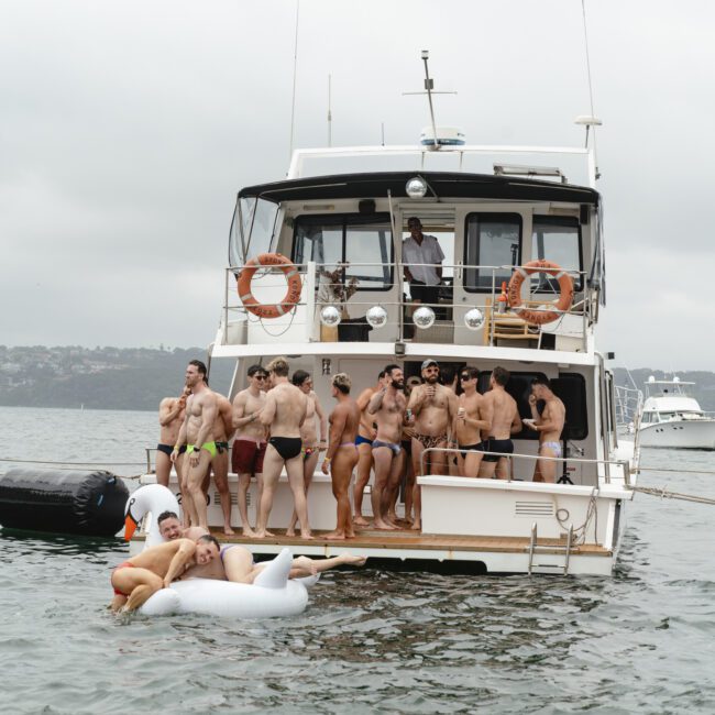 A group of people in swimsuits stand on the deck of a yacht, while one person floats nearby on an inflatable swan. The scene is set on a cloudy day with another boat visible in the background.