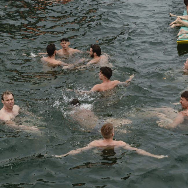 A group of people swimming and splashing in a body of water, with some floating on an inflatable object. The scene is lively and the water appears to be a natural outdoor setting.