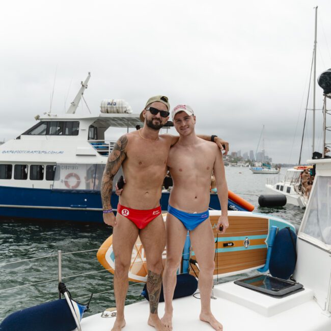 Two men in swim briefs stand arm in arm on a boat deck. One wears red, the other blue. A larger boat and a city skyline are visible in the background under a cloudy sky.