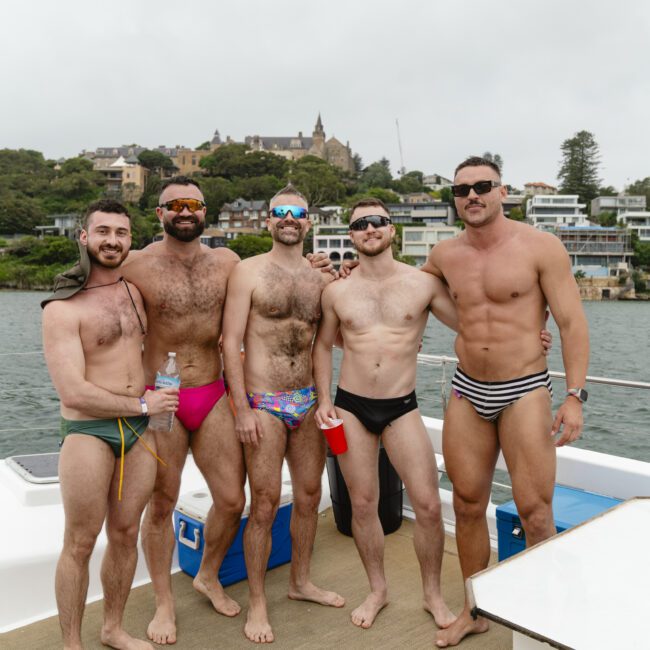 Five men in colorful swimwear stand together on a boat, smiling at the camera. In the background, there are waterfront buildings and greenery under a cloudy sky.